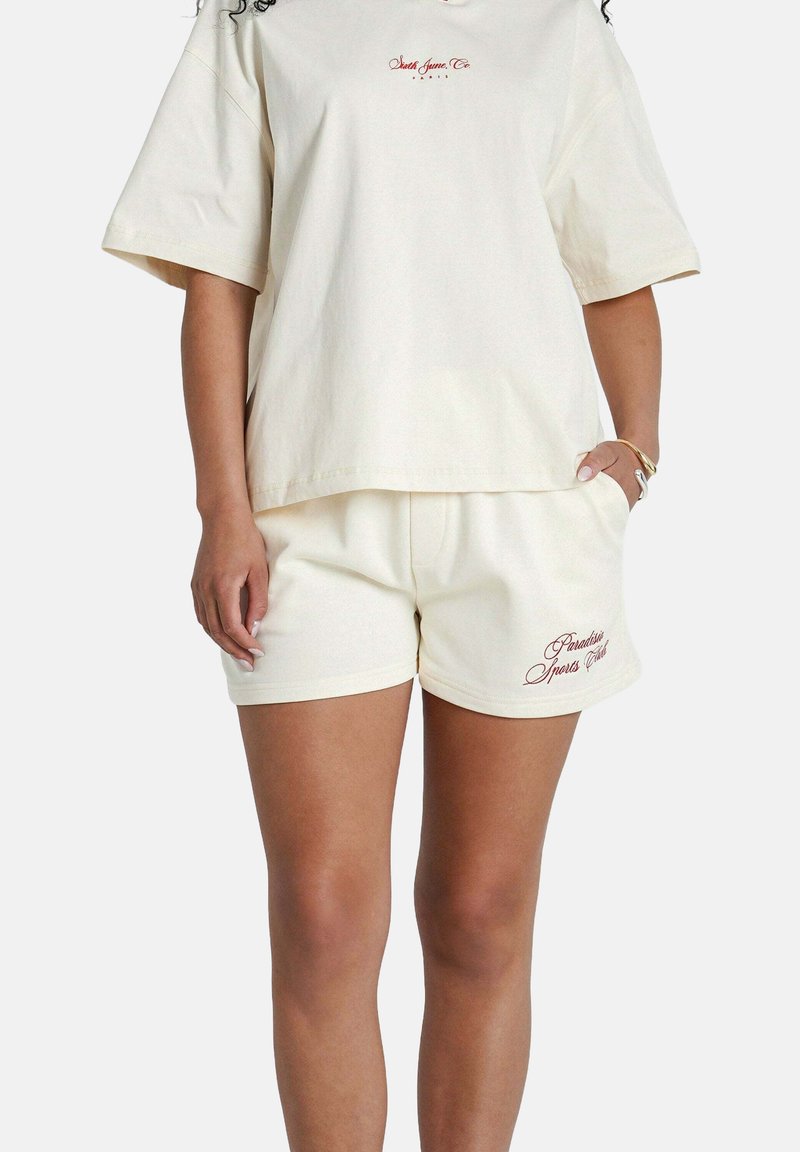 Maglietta in cotone color crema con maniche corte e logo ricamato. Abbinata a pantaloni corti coordinati con tasche laterali e cuciture decorative.