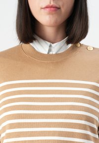 Beiger Pullover mit weißen horizontalen Streifen, weichem Stricktextur, rundem Ausschnitt und goldenen Knopf-Akzenten an der Schulter, getragen über einem Hemd mit Kragen.