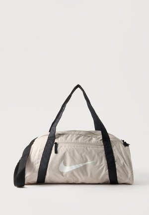 Svetlobéžová športová taška Nike s čiernymi rukoväťami, predným vreckom na zips a bielym logom Swoosh na jednotnom svetlom pozadí.