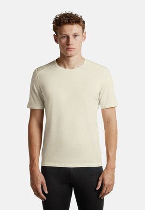 Joven con cabello rizado de pie mirando hacia adelante, vistiendo una camiseta lisa beige de manga corta y pantalones negros sobre un fondo blanco.