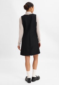 Schwarzes ärmelloses Kleid aus strukturiertem Stoff, mit einem Knopfdetail am Rücken und einem ausgestellten Saum, getragen über einem langen beigen Oberteil und kombiniert mit schwarzen Schuhen.
