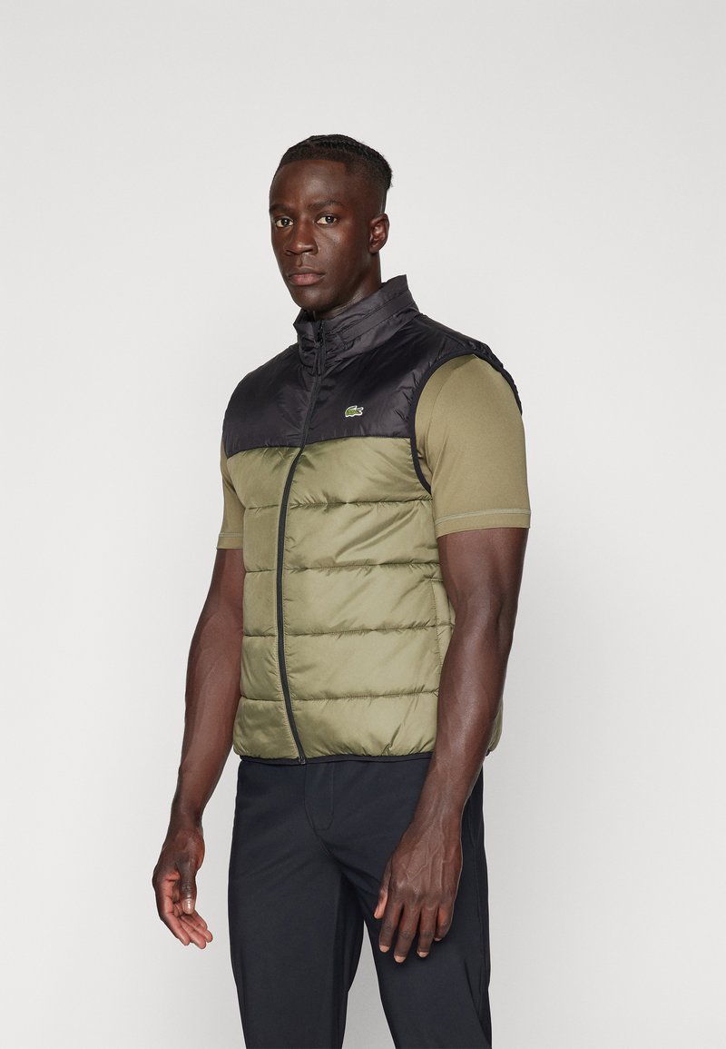 Gilet sans manches isolé avec une partie inférieure matelassée en vert olive et une partie supérieure noire. Présente un col montant et une fermeture éclair à l'avant.