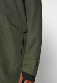 Giacca impermeabile verde con finitura testurizzata, caratterizzata da polsini aderenti, tasca con zip laterale e tasca a patta sul braccio.