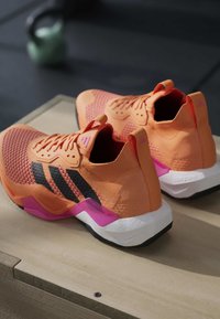 Chaussures de sport orange et rose avec des rayures noires posées sur une surface en bois, un kettlebell vert flou en arrière-plan.