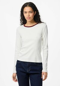 Kvinna med lockigt mörkt hår som bär en vit långärmad skjorta med mörkröd krage och mörkblå jeans, stående mot en vit bakgrund.