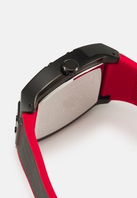 Montre noire en métal avec un boîtier rectangulaire, un bracelet en silicone rouge, une surface texturée et un bouton latéral proéminent. Dos en argent avec des détails gravés.
