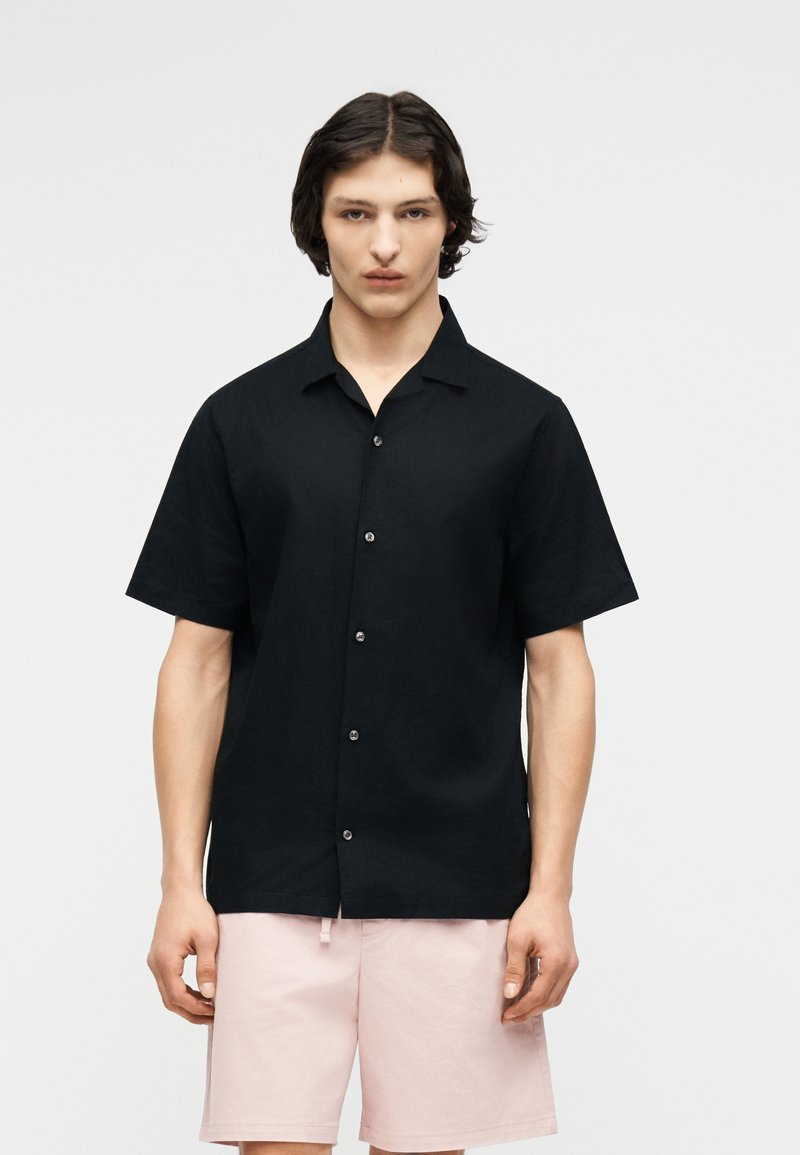 Νέος άνδρας που φοράει μαύρο πουκάμισο με κοντά μανίκια και ροζ σορτς, στέκεται μπροστά σε απλό φωτεινό φόντο.