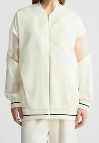 Bílá bomberka s průhlednými síťovými panely, zipovým zapínáním a kontrastními černými pruhy na manžetách a lemu. Obsahuje geometrické designové prvky.