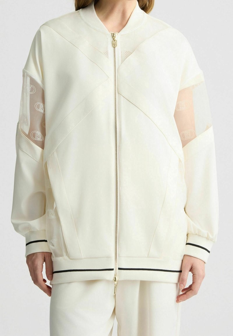 Bílá bomberka s průhlednými síťovými panely, zipovým zapínáním a kontrastními černými pruhy na manžetách a lemu. Obsahuje geometrické designové prvky.