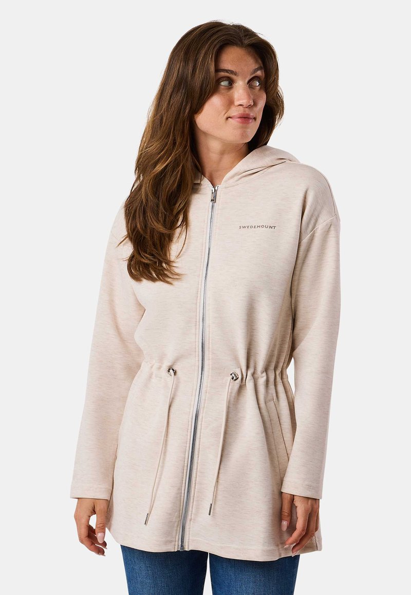 Veste zippée à capuche beige avec taille à cordon et manches longues. Présente une texture subtile et un petit logo sur la poitrine gauche.