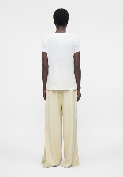 Person with short curly hair wearing a white short-sleeve shirt and wide beige pants, standing facing away against a plain background.