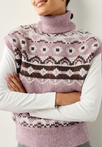 Gebreid vest met een hoge col, met roze, witte en bruine geometrische patronen. Draagt over een wit top met lange mouwen. Zachte textuur.