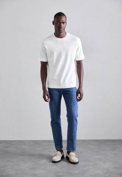 Chemise blanche texturée à manches courtes, jean en denim bleu et mocassins clairs avec des accents métalliques. Design simple, coupe décontractée.
