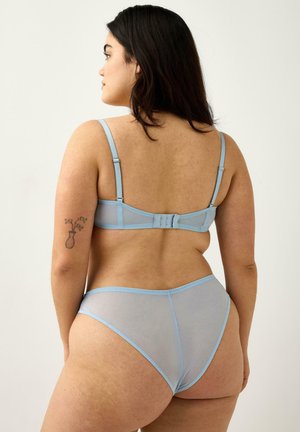 Donna con lunghi capelli scuri che indossa un reggiseno azzurro chiaro trasparente e slip coordinati, vista di schiena su sfondo chiaro uniforme.