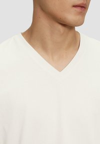 Weißes V-Ausschnitt-T-Shirt aus Baumwolle mit weicher Textur und dezentem Prägungslogo auf der linken Brust. Nahaufnahme des Ausschnitts und der Schulter.