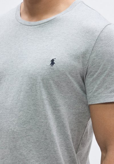 Pelēka īsroku t-krekla ar apaļu kakla izgriezumu, izgatavota no mīksta kokvilnas materiāla, ar tumši zilu polo logo kreisajā krūšu daļā.