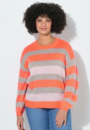 Kvinde med krøllet hår iført en stribet sweater i lys orange, grå og pink samt blå jeans, stående foran en hvid baggrund.