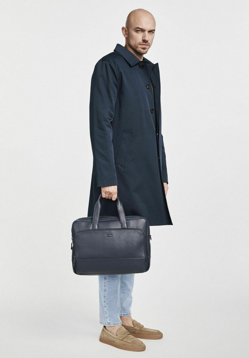 Ciemnoniebieski, wodoodporny płaszcz z dużymi guzikami; trzyma skórzaną torbę na laptopa o teksturowanej powierzchni. Jasne dżinsy i brązowe loafersy dopełniają wygląd.