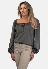 Szara, jedwabna bluzka z kwadratowym dekoltem, długimi bufkami i wiązaniem na sznurku z przodu, w zestawieniu z beżowymi dopasowanymi spodniami.