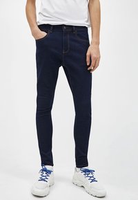 Mörkblå slim-fit jeans med fem fickor, orange sömmar och en mjuk textur. Kombinerade med vita sneakers med blå snören.