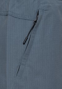 Nærbillede af en blå lynlåslomme med en sort trækflig på et tekstureret materiale til udendørs- eller aktivtøj.