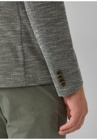 Grijze textured blazer met een enkele knoopsluiting en vier bruine knopen op de manchetten. Gedragen over lichtgroene broeken, die een getailleerde pasvorm laten zien.