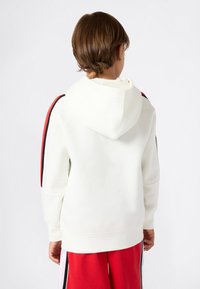 Witte hoodie met een capuchon, voorzien van rode en zwarte zijstrepen, gemaakt van zachte stof. Gedragen met rode trainingsbroek, tentoongesteld vanuit de achterkant.