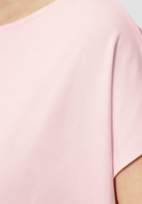 Jasnoróżowa bluzka z krótkim rękawem o gładkiej strukturze, okrągłym dekolcie, luźnym kroju i minimalnymi detalami przeszycia wzdłuż dołu.