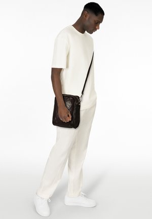 Borsa a tracolla in pelle marrone con cerniera, indossata da una persona con t-shirt bianca e pantaloni, in piedi davanti a uno sfondo bianco uniforme.