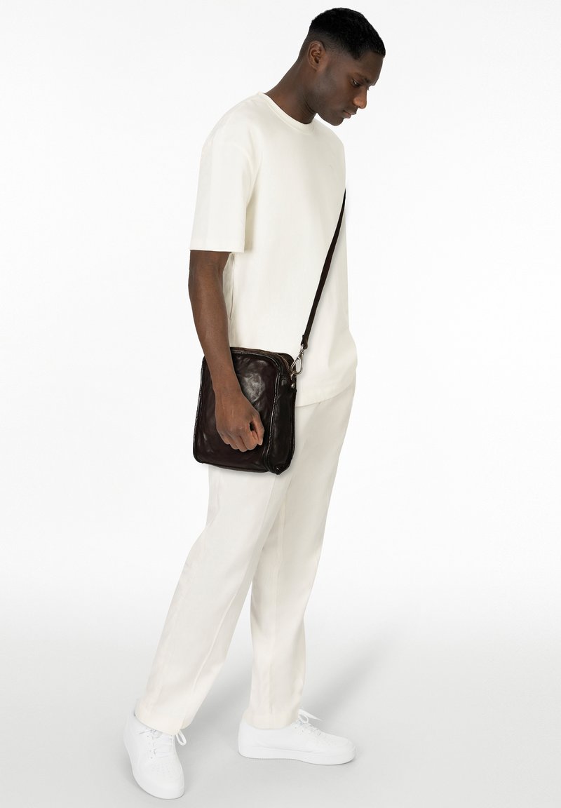 Borsa a tracolla in pelle marrone con cerniera, indossata da una persona con t-shirt bianca e pantaloni, in piedi davanti a uno sfondo bianco uniforme.