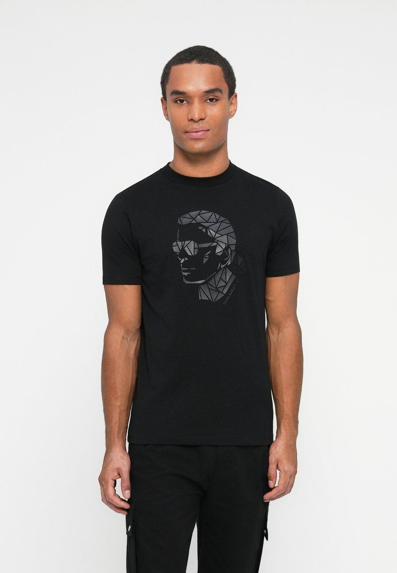 Hombre vestido con una camiseta negra con un diseño de cara geométrica en impresión negra brillante, combinada con pantalones cargo negros, de pie frente a un fondo blanco.