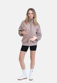 Světle hnědá mikina na půl zip s velkou přední kapsou, v kombinaci s černými šortkami a bílými teniskami, na jednoduchém pozadí.