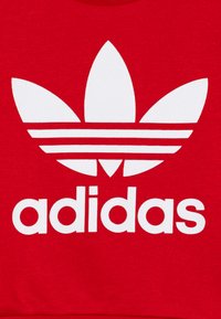 Rdeča bombažna majica s kapuco z velikim belim logotipom trefoil Adidas in imenom blagovne znamke v krepki pisavi. Gladka tekstura s oblikovanjem okroglega izreza.
