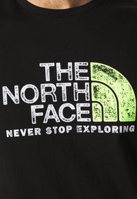 Černé tričko s opotřebovaným bílým a neonově zeleným logem „THE NORTH FACE“ a nápisem „NEVER STOP EXPLORING“ v tučném písmu.