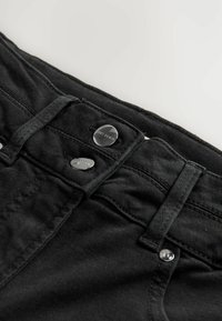 Jeans de mezclilla negra con dos botones metálicos en la cintura, costuras distintivas y una textura suave. Vista en primer plano de la parte superior.