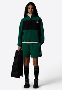 Veste coupe-vent verte et noire avec poches zippées, associée à un short vert. Doudoune noire tenue, chaussettes noires montantes et bottes à lacets.