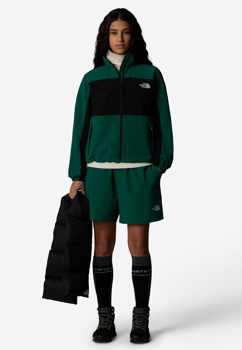 Veste coupe-vent verte et noire avec poches zippées, associée à un short vert. Doudoune noire tenue, chaussettes noires montantes et bottes à lacets.