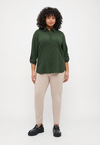 Tmavě zelená košile na knoflíky s texturovanou tkaninou, ohrnuté rukávy, spárovaná s béžovými slim-fit kalhotami a černými robustními botami s hroty.
