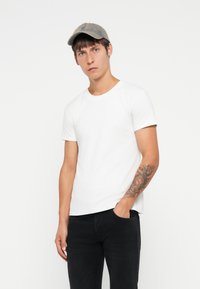 T-shirt bianca in cotone a maniche corte, abbinata a jeans neri. Un cappellino grigio riposa sulla testa e sul braccio sinistro c'è un tatuaggio.