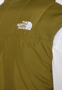 Olivgrön väst med quiltad textur, prydd med en vit broderad logotyp. Designen inkluderar en hög krage och ärmlös skärning, lagd över en vit skjorta.