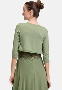 Zielony krótkotrwały top z rękawami trzy czwarte, z koronkowym akcentem na plecach, w zestawieniu z luźną zieloną sukienką. Miękka faktura materiału.