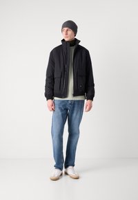 Jachetă bomber neagră cu două buzunare frontale, purtată cu un tricou verde deschis, blugi albaștri și adidași albi. Model care poartă o căciulă gri.