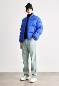 Karališkai mėlyna pūkinė striukė su užtrauktuku, juodas megztinis su aukštu kaklu, šviesiai mėlyni džinsai ir balti sportbačiai. Juodas megztinis su "HUGO" logotipu.