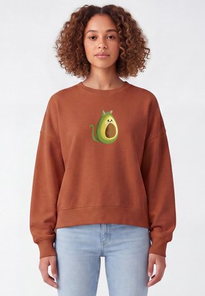 Vrouw met krullend haar die een bruine trui draagt met een cartoon avocado in de vorm van een kat, gecombineerd met lichtblauwe spijkerbroek.