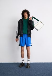 Black zip-up jacket with patterned design, green polo shirt, blue shorts, black shoes with thick soles, and a white shoulder bag with green accents.