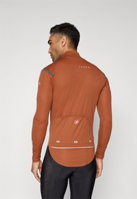 Maillot de cyclisme à manches longues en tissu marron avec une texture lisse, présentant des accents réfléchissants et des poches arrière. Inclut un détail de marque à l'arrière.