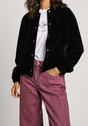 Nainen, joka pukeutunut mustaan samettiseen napilliseen takkiin, valkoiseen graafiseen T-paitaan ja vaaleanpunaiseen korkeavyötäröiseen farkkuihin, joihin hänellä on käsi taskussa.