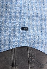 Camicia a maniche corte di tessuto azzurro chiaro con un motivo geometrico in blu scuro. Etichetta visibile vicino all'orlo. Abbinata a jeans grigi scuri.