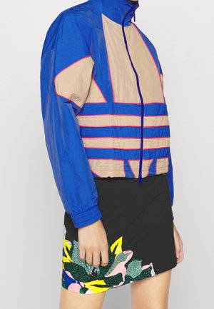 Personne portant une veste zippée bleue et beige avec une bordure rose et une jupe noire avec des motifs floraux colorés près de l'ourlet.