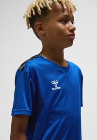 Blå sporttröja av slätt tyg med korta ärmar, V-ringning och svarta ränder på axlarna. Logo visas på bröstet.